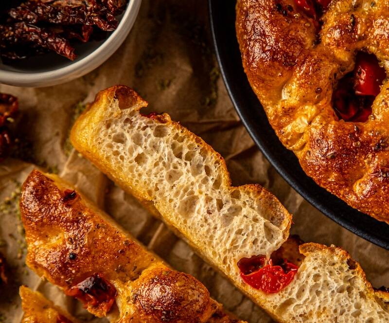 Porciones de focaccia de tomate y orégano, junto a media focaccia en plato y un cuenco con tomates enteros, elaborada de forma artesanal con aceite de oliva virgen extra.