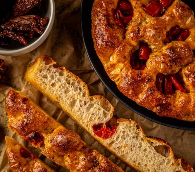 Porciones de focaccia de tomate y orégano, junto a media focaccia en plato y un cuenco con tomates enteros, elaborada de forma artesanal con aceite de oliva virgen extra.