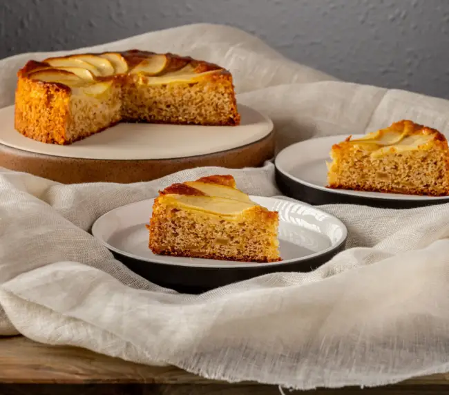 Bizcocho de manzana artesanal de Andrea Forte Panadería-Pastelería cortado en porciones, servido en plato con dos platos pequeños que contienen una porción de tarta cada uno.