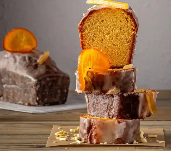 Porciones de cake de cítricos apiladas en forma de torre en primer plano, con bizcocho de limón y naranja glaseado y decorado con fruta confitada, y el pastel entero al fondo.