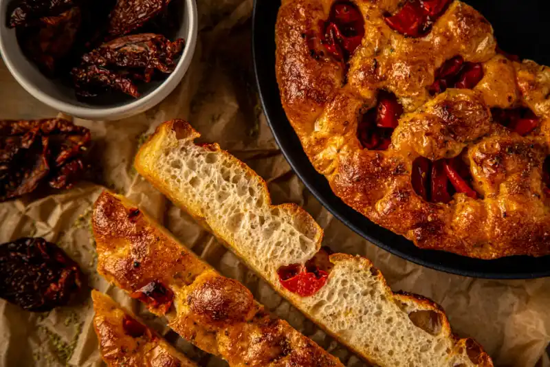 Porciones de focaccia de tomate y orégano, junto a media focaccia en plato y un cuenco con tomates enteros, elaborada de forma artesanal con aceite de oliva virgen extra.