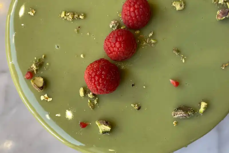 Tarta de color verde claro decorada con tres frambuesas rojas y trozos de pistacho.