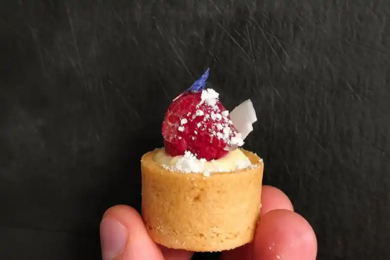 Mano sosteniendo un pequeño pastel tipo mignardise con una frambuesa como decoración.